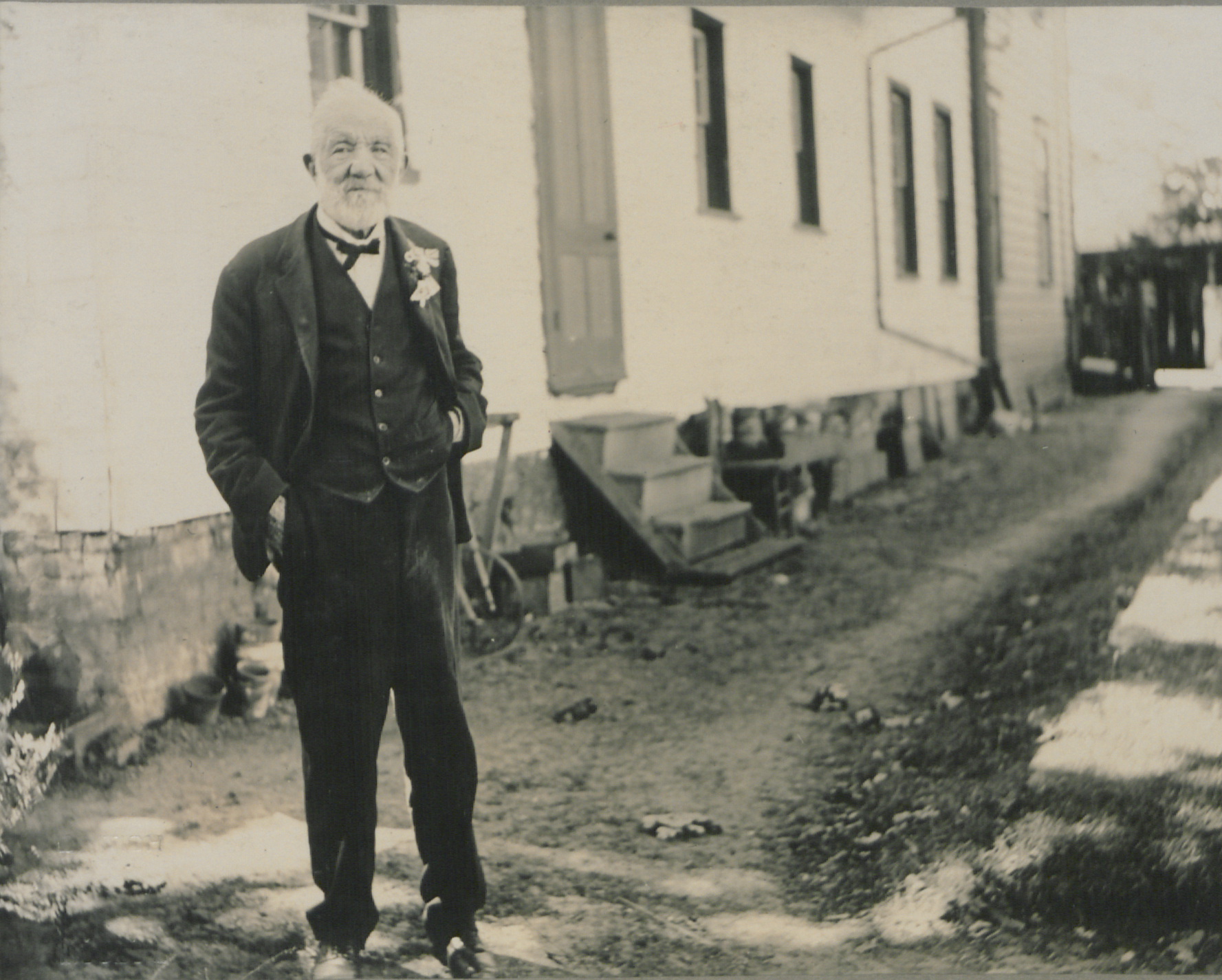 Old man in a suit stands outside a house on a dirt road.