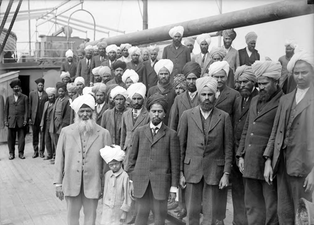 Forty men in suits and turbans stand on the deck of a ship.