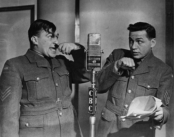 Two men in military uniforms stand by a broadcast microphone. One holds a small comb under his nose.