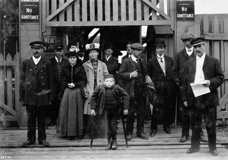 Eight men, two women, and a boy stand in front of a tall wooden gate.