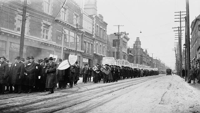 Men march down street carrying signs. Many wear overcoats and hats. Some carry musical instruments.