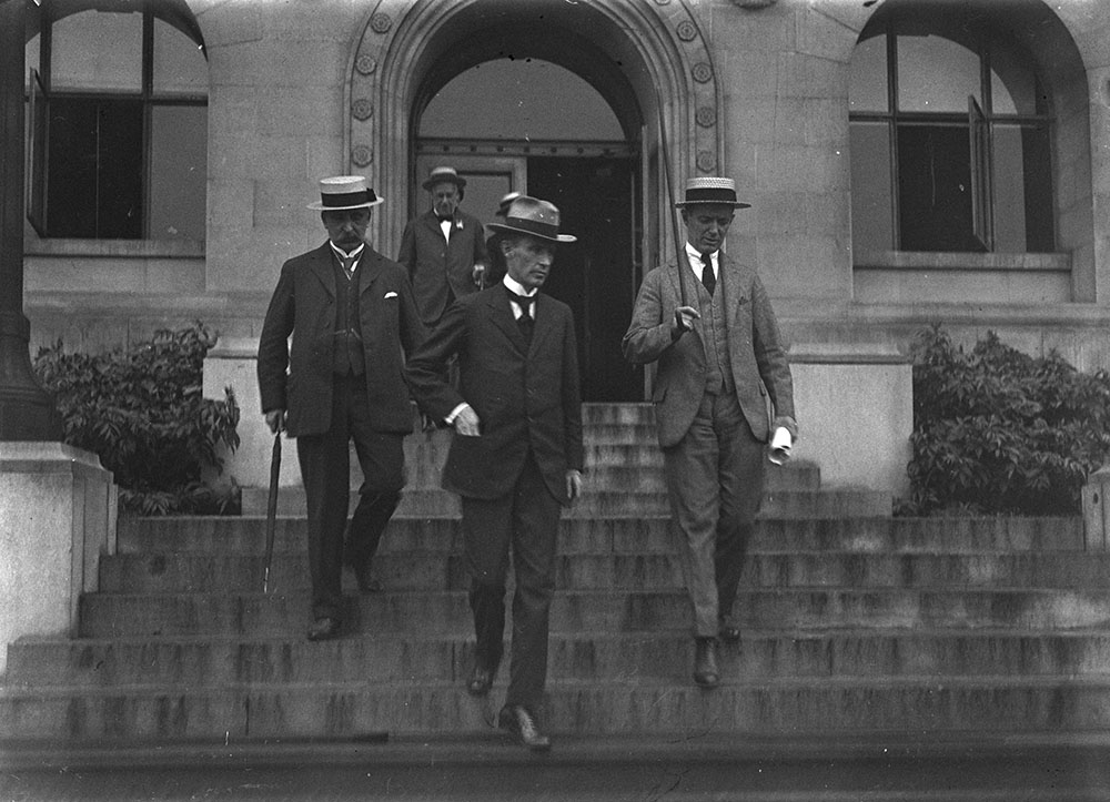 Four men in suits and flat straw boater hats descend stone steps.