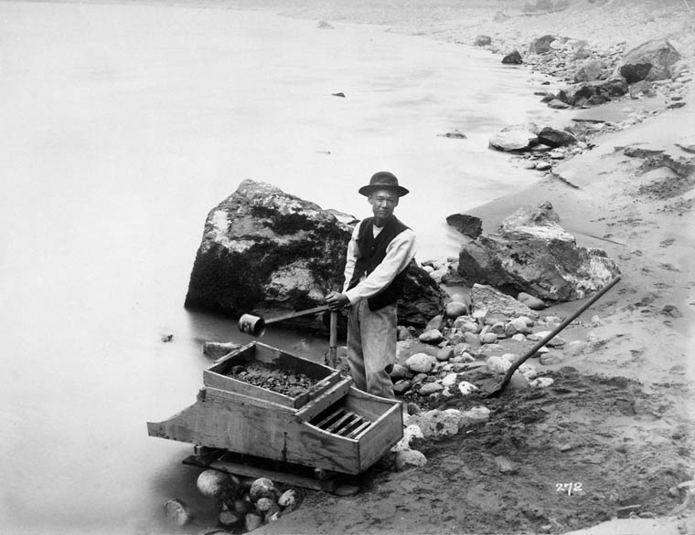 Man washing gold at riverside using a wooden box with a metal grate.