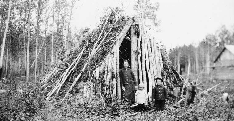 A woman and two children stand in the doorway of a lean-to in a wooded area.