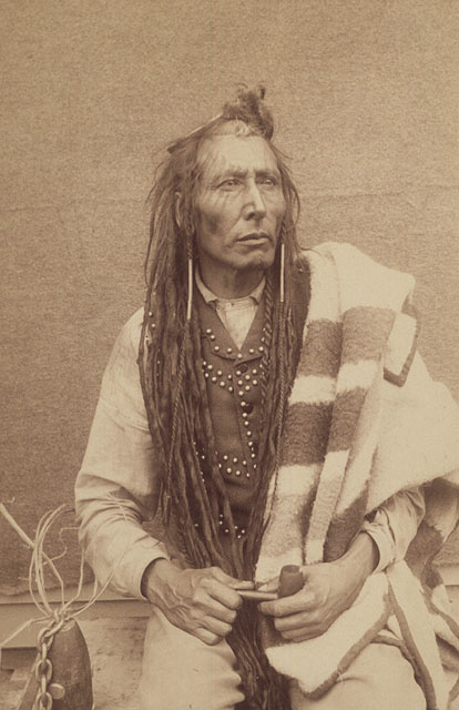 A man with long braids and a blanket over his shoulder holds a smoking pipe and looks pensive.