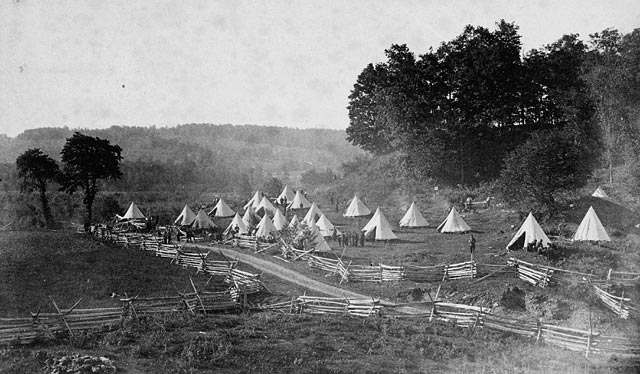 A dozen or so tents pitched in a field with a few trees surrounded by a simple wooden fence.