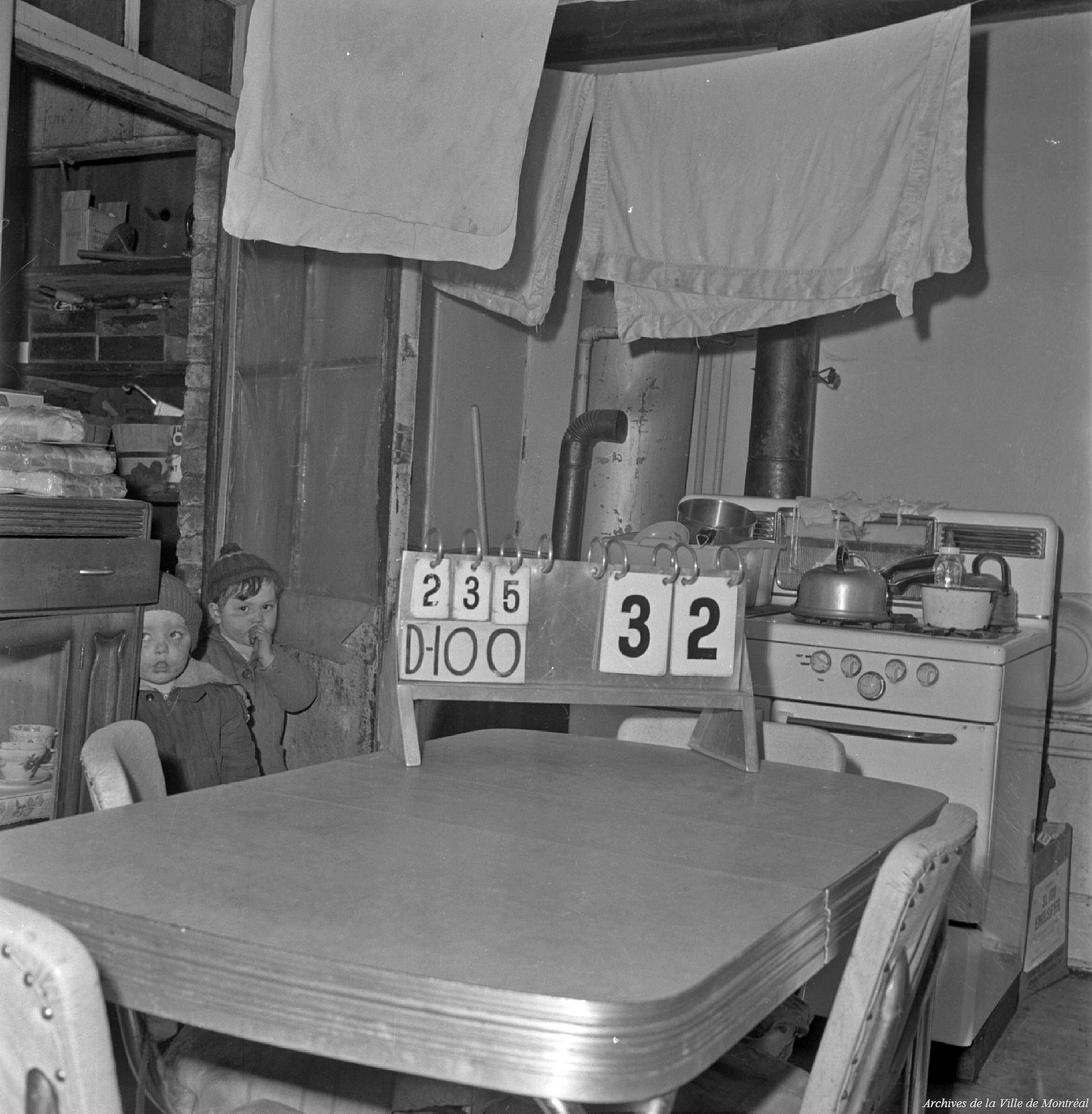 Two children stand in a kitchen with blankets hanging on laundry lines.