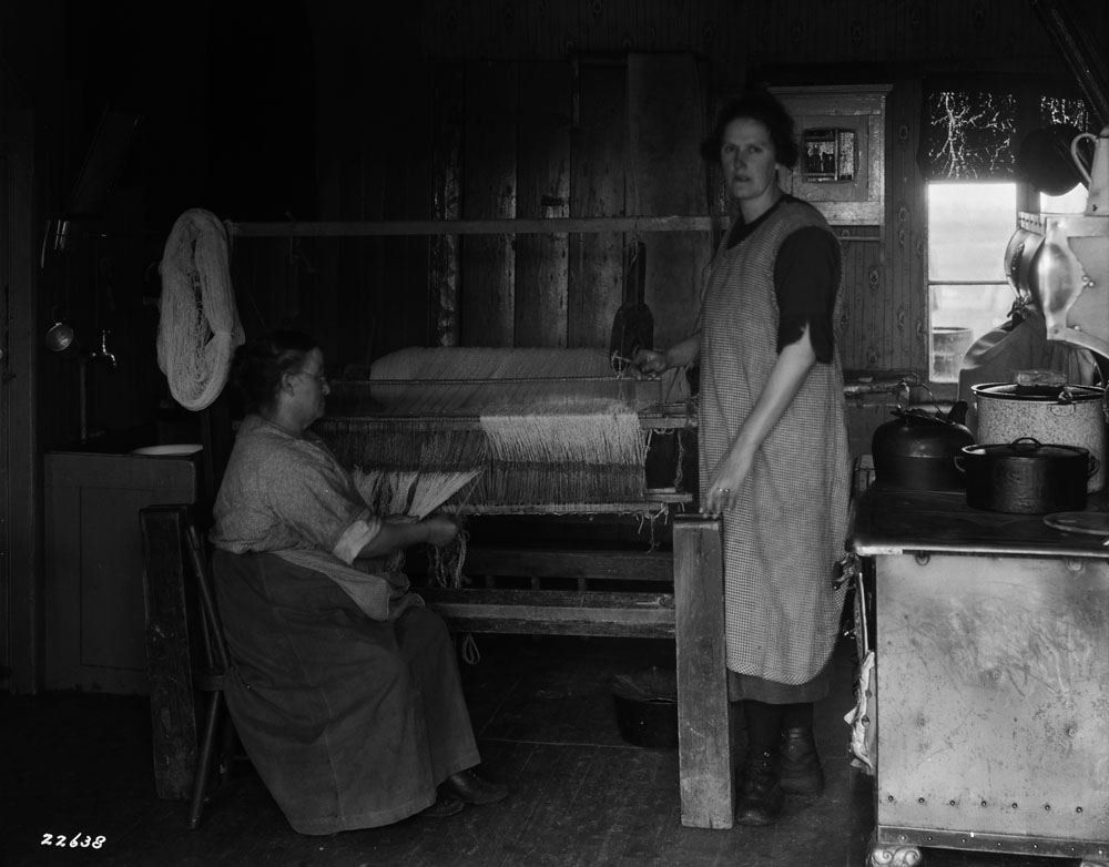 Two women work on a loom in a cramped kitchen.