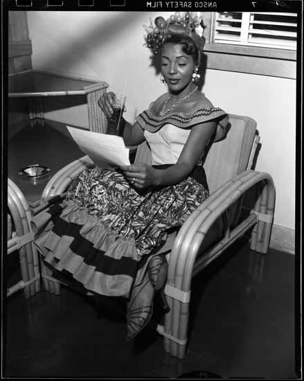 A young black woman reviews a document, drink in hand. She wears a Carmen Miranda–esque costume.