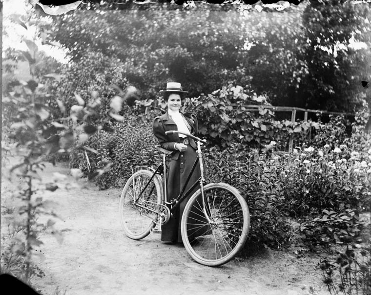 A woman in a Victorian dress poses with a bicycle in a garden.