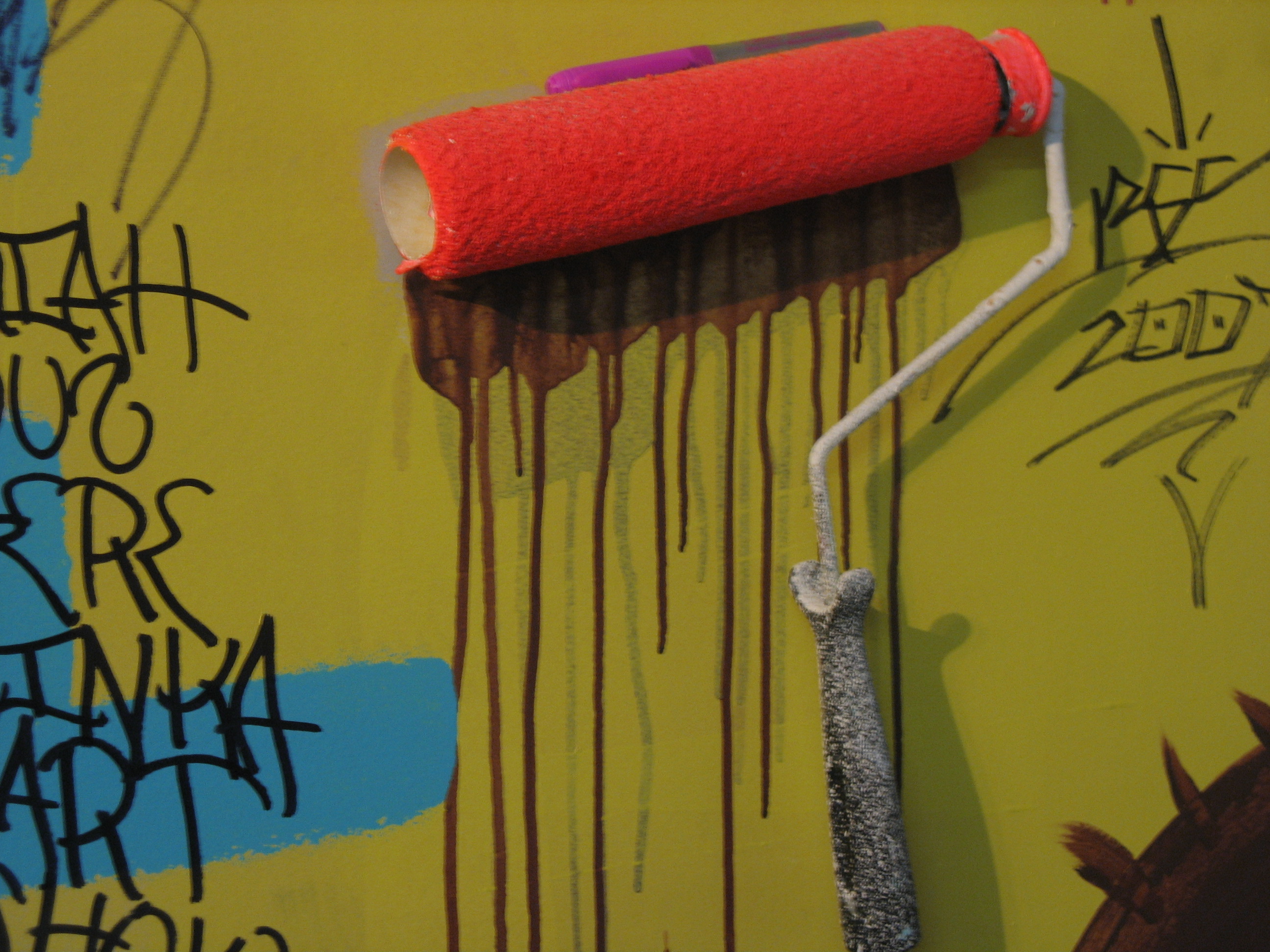 An orange paint roller is pushed against a wall. Paint drips down the wall beneath it.