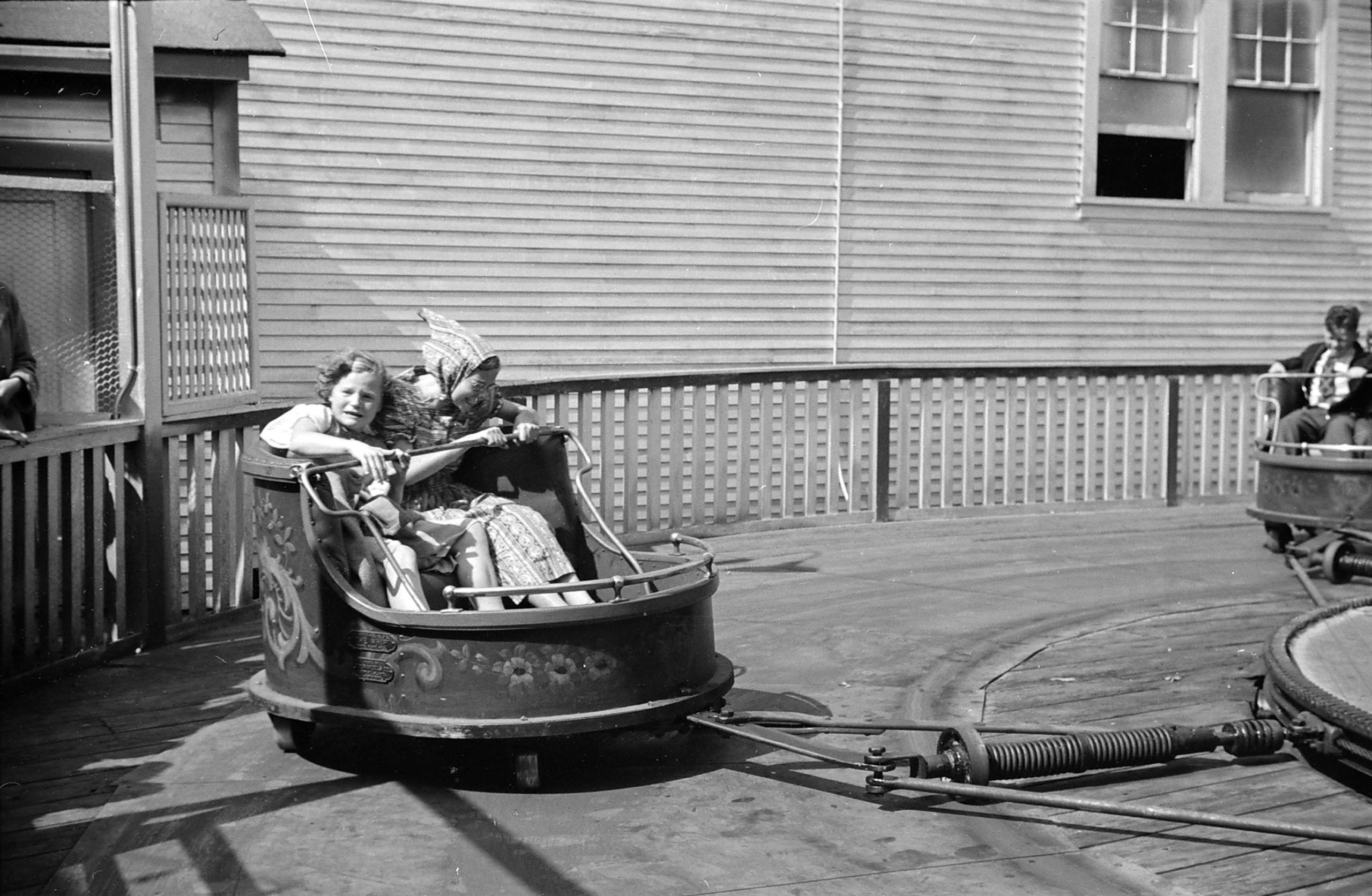 Two girls sit in a fair ride car swinging in a circle. One looks thrilled, while the other scared.