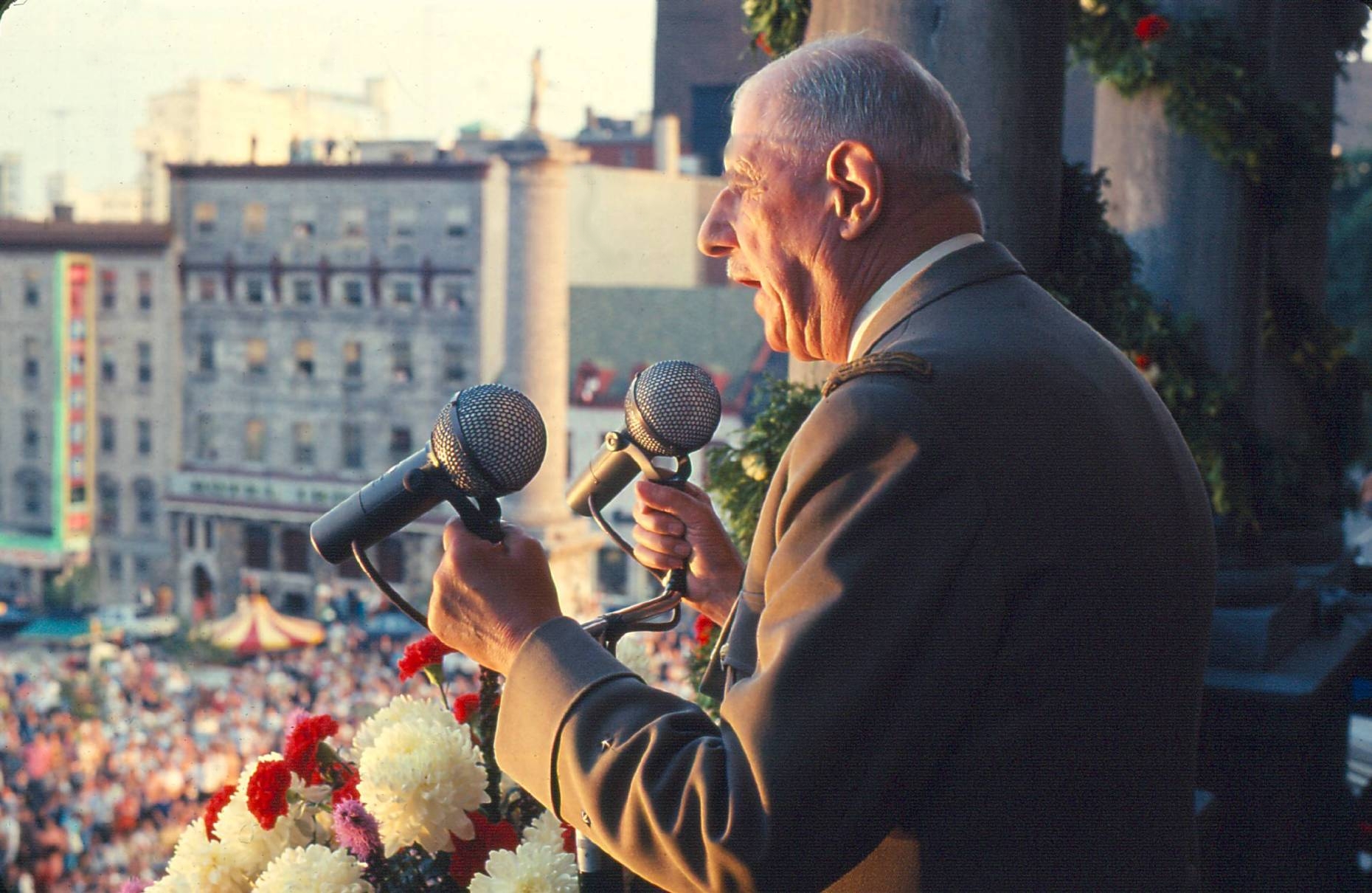 An older man on a balcony grips two microphones and addresses an unseen crowd on the street.