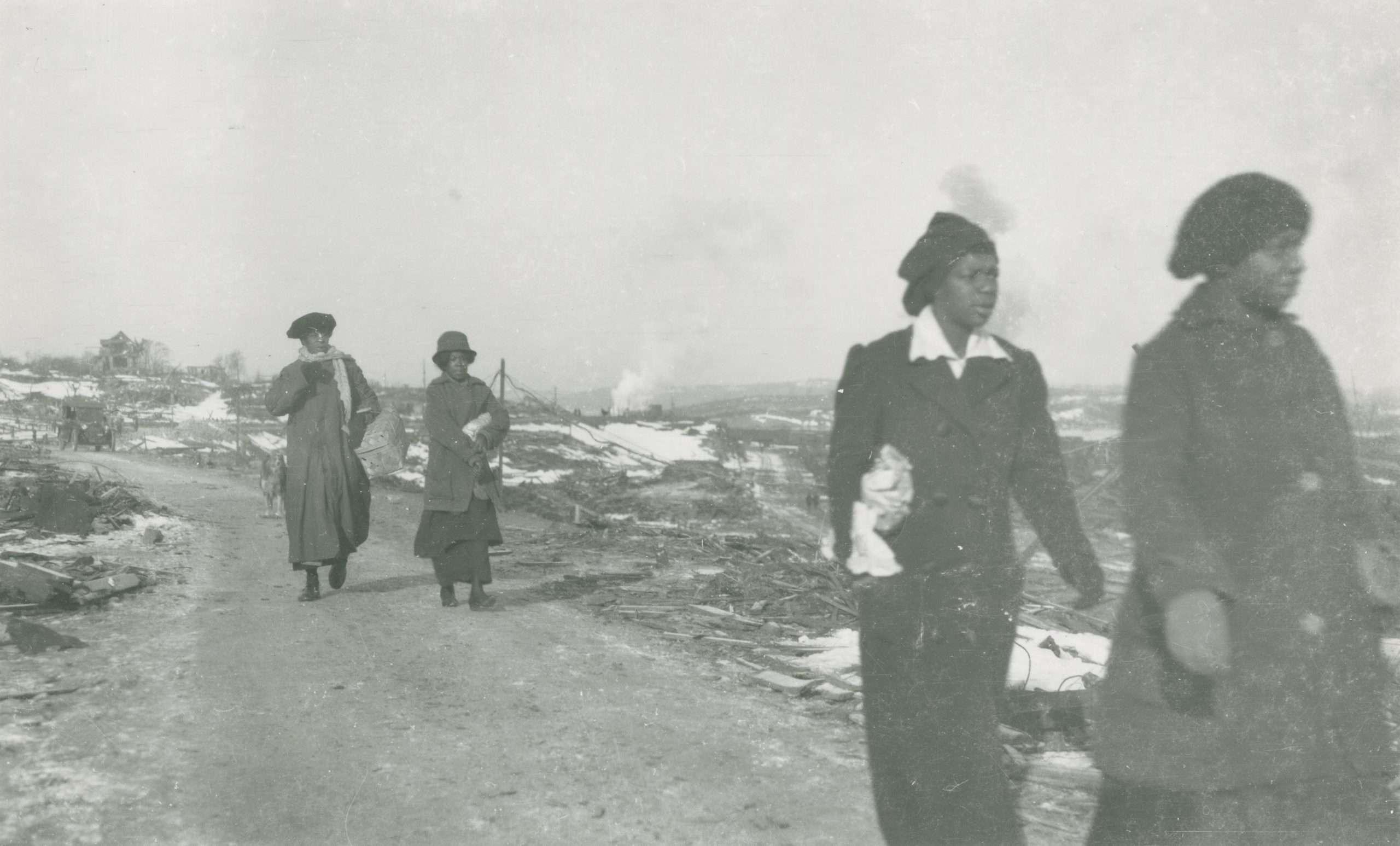 Four Black women in overcoats walk down a dirt road that runs through barren land.
