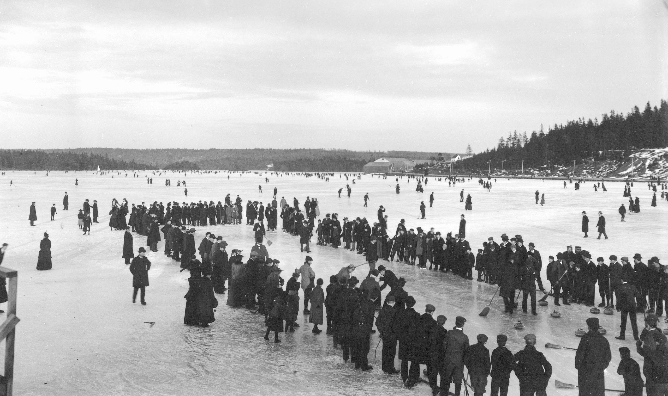 People line a curling rink on a frozen lake. People skate and play in the snow in the background.