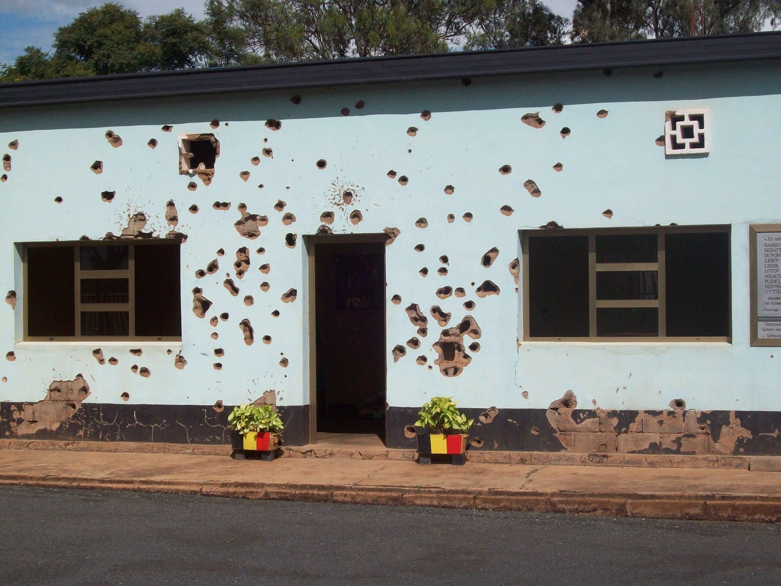 A small building riddled with bullet holes. Two potted plants outside the door bear Belgian colours.