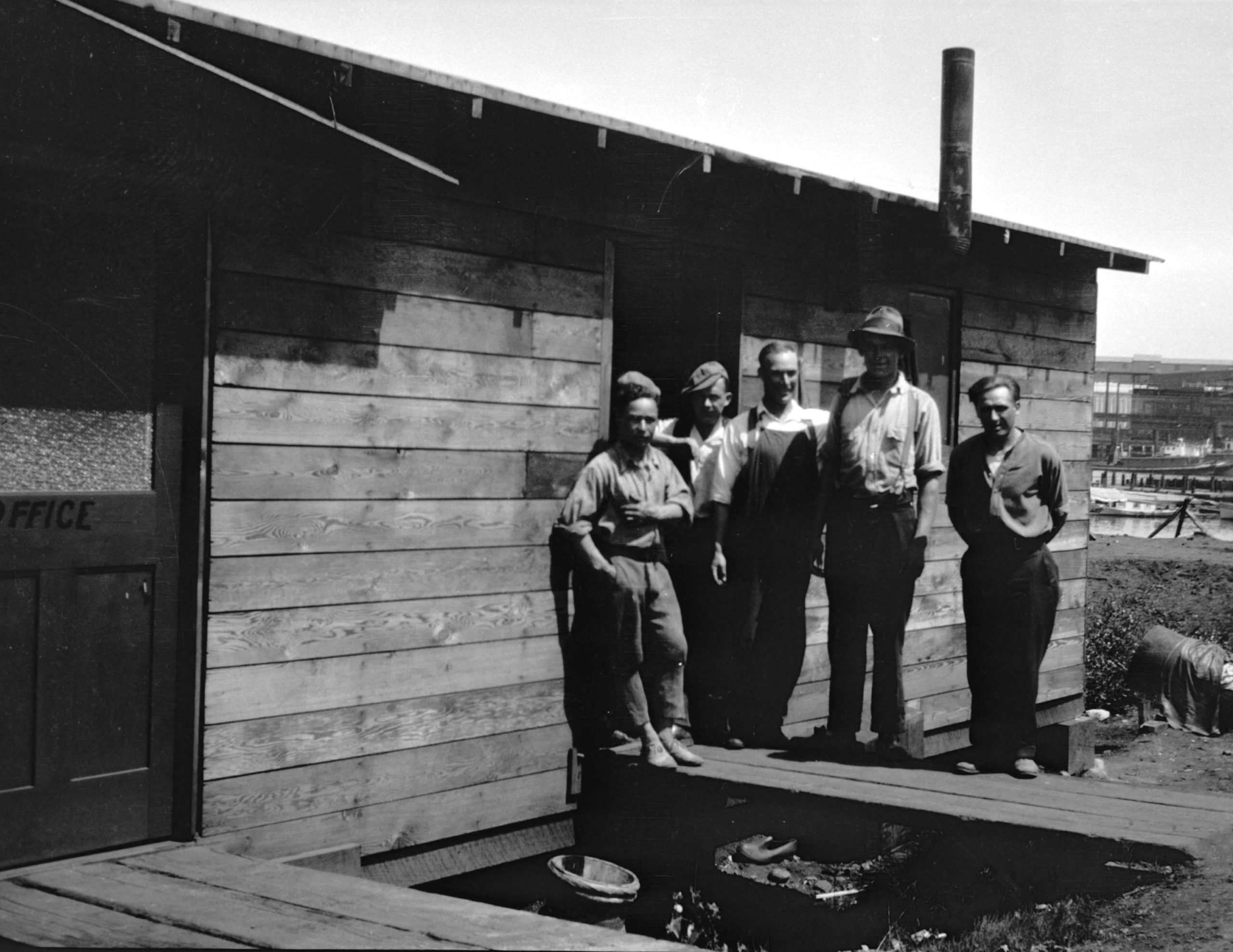 Five men stand outside of a wooden shack.