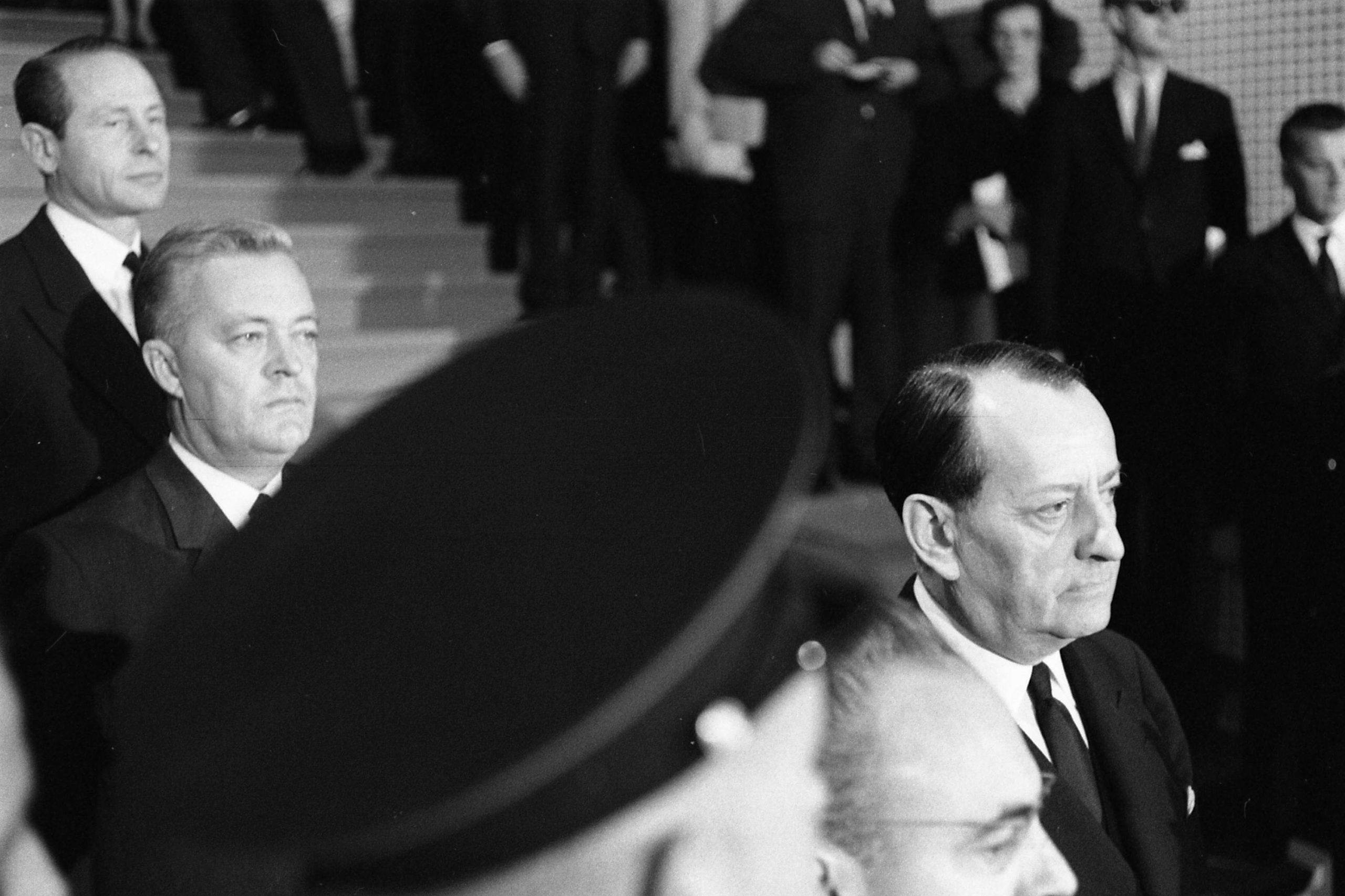 Three men in suits descend a staircase. A military officer&#039;s hat obscures most of the shot.