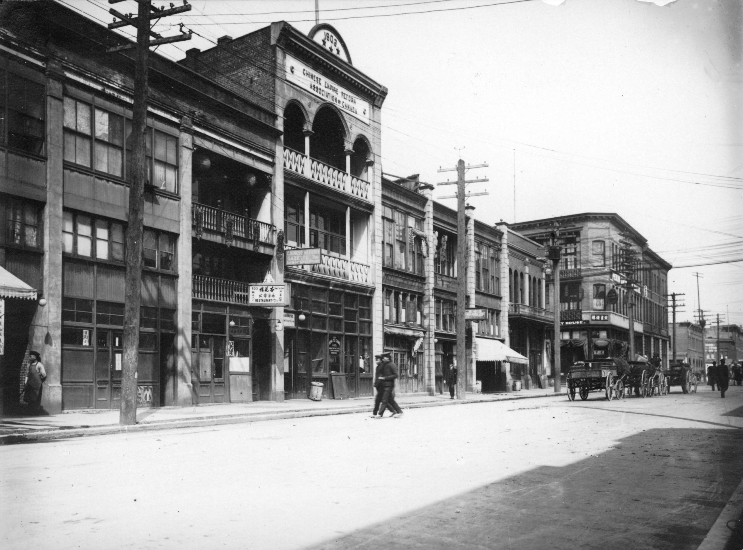 A city street in 1907. A few carriages and pedestrians go down the street past storefronts.