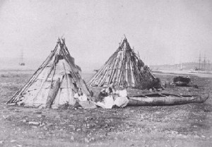 People sit outside of two teepees. A canoe sits beside them.