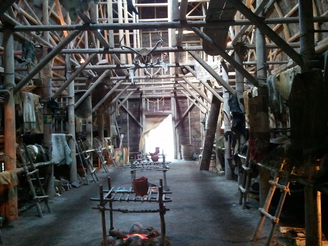 Photo of the inside of a long building with log rafters and replica wooden grills over small fires.
