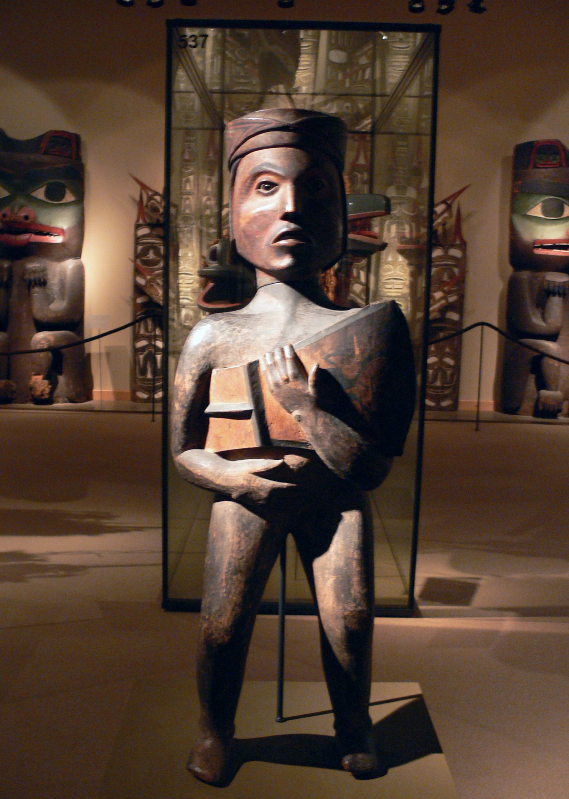 Metal statue of a man holding a copper shield in a museum. Behind the statue are Indigenous carvings.