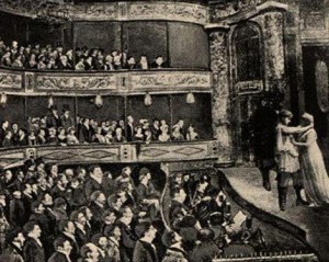 People in suits and fancy clothes sit in the audience and watch actors on the stage.