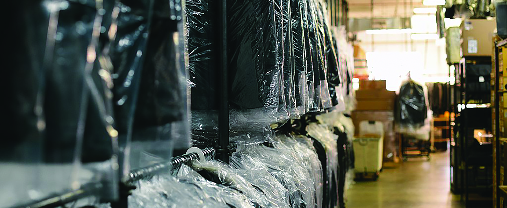 View of interior of dry-cleaning business. On the left are racks of clothes covered in plastic. On the right are shelves holding boxes.