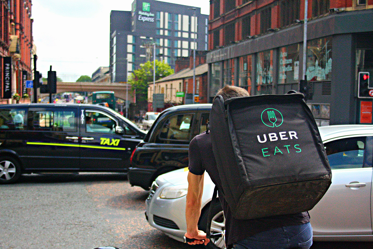 A photo shows the rear view of an Uber Eats’ food delivery driver with a backpack, cycling along a busy Oxford Road in Manchester, England.