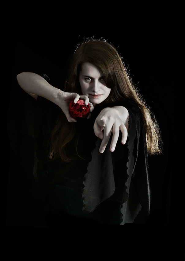 A woman stares intensly into the camera. Her hands are stretched in front of her body like a creature and her dark clothes blend in with the dark background.