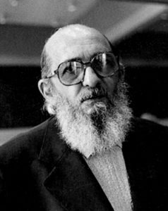 Photo portrait of a balding middle-aged man with a medium-length white beard, wearing large glasses and a suit.