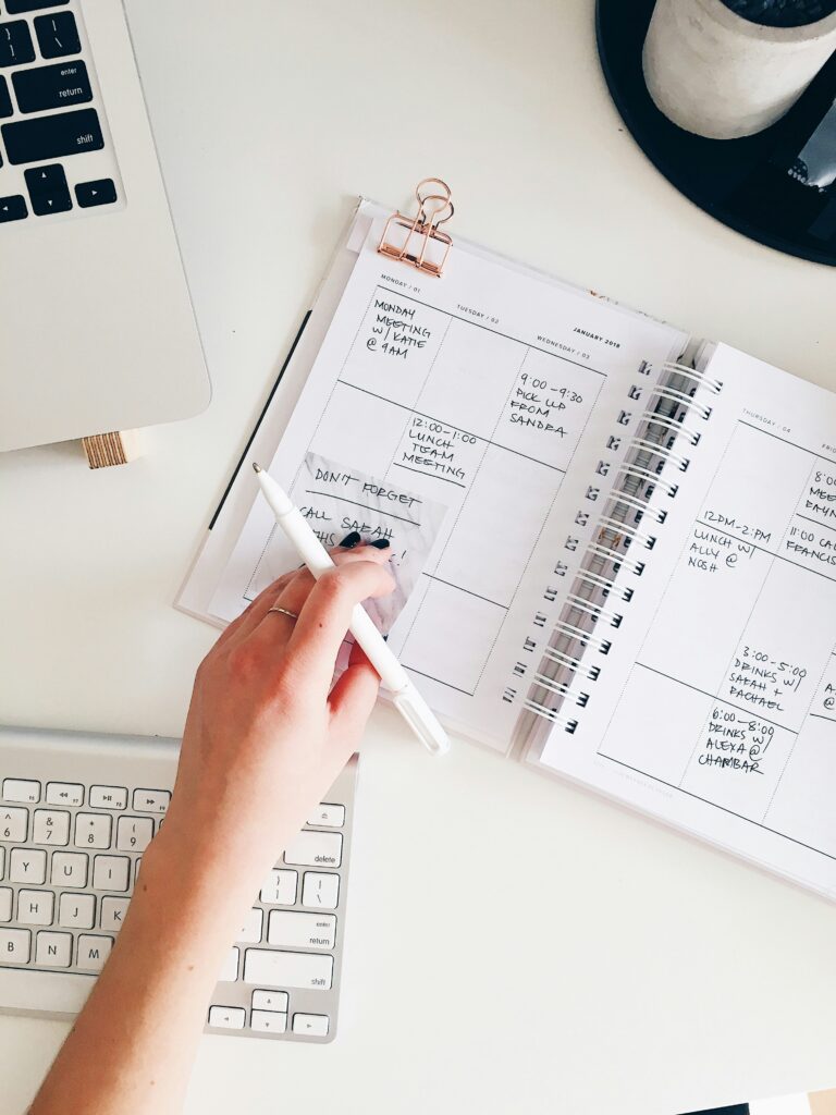 A hand holds a pen and touches an open planner filled with events.