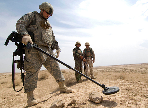Photograph shows a soldier with the metal detector in one hand.