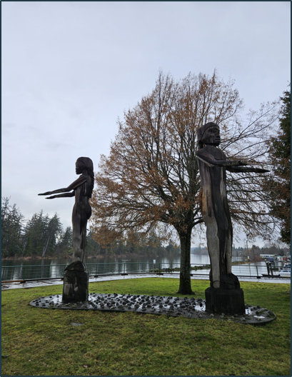 Two tall, wooden welcome figures facing away from each other. One is male and the other is female. Their arms are stretched out in front with palms up.