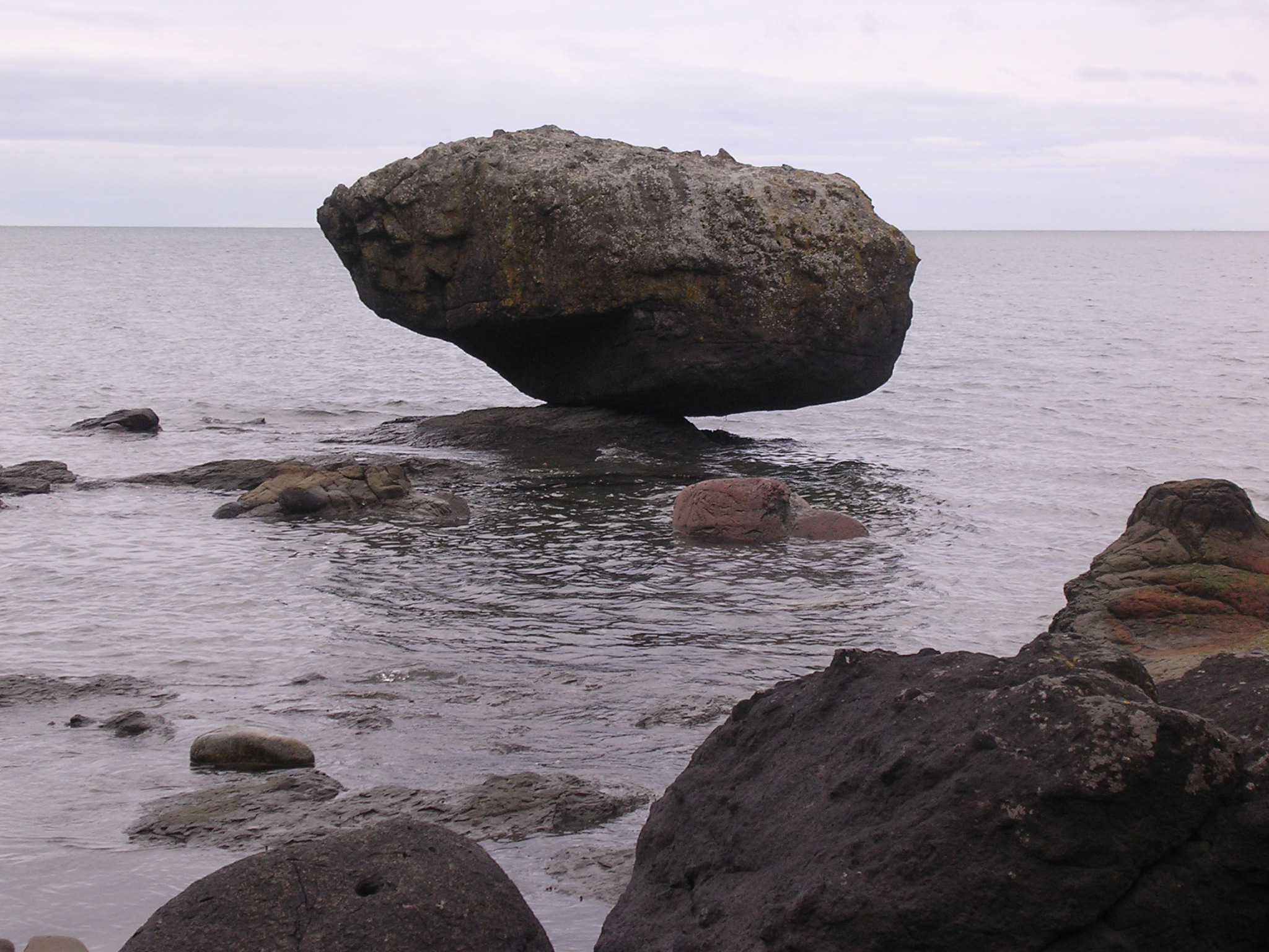 A massive boulder balances on top of a small rock in the ocean.