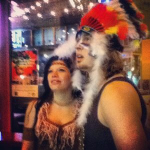 A man and woman pose in costumes that represent stereotypes about Indigenous Peoples, including a war bonnet and tan clothing with tassels.